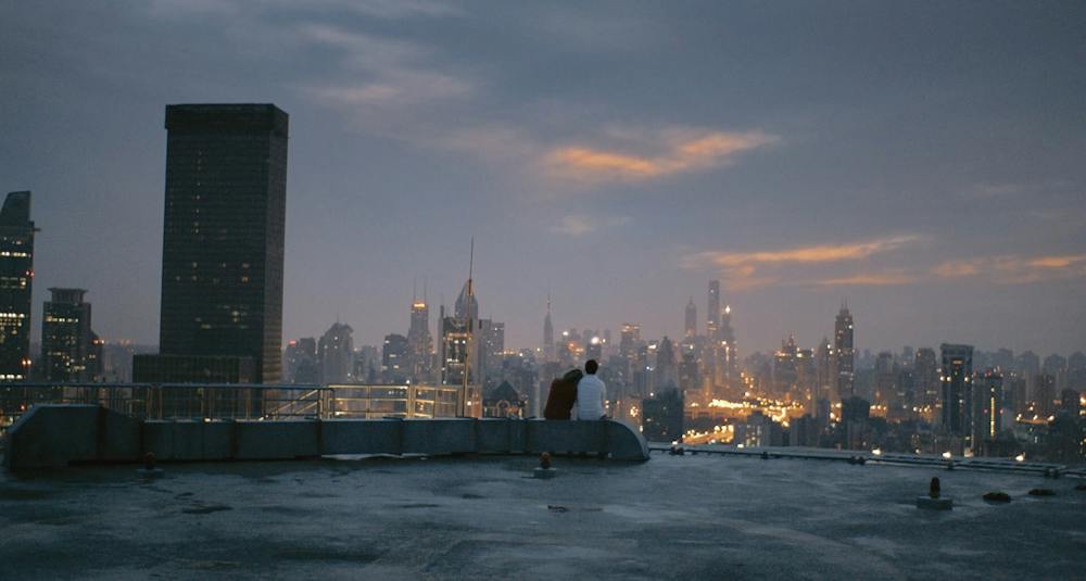Theodore and Amy seated together on a rooftop above the city at dawn